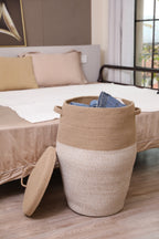 Jute and white laundry hamper beside a bed holding folded laundry, lid resting against it